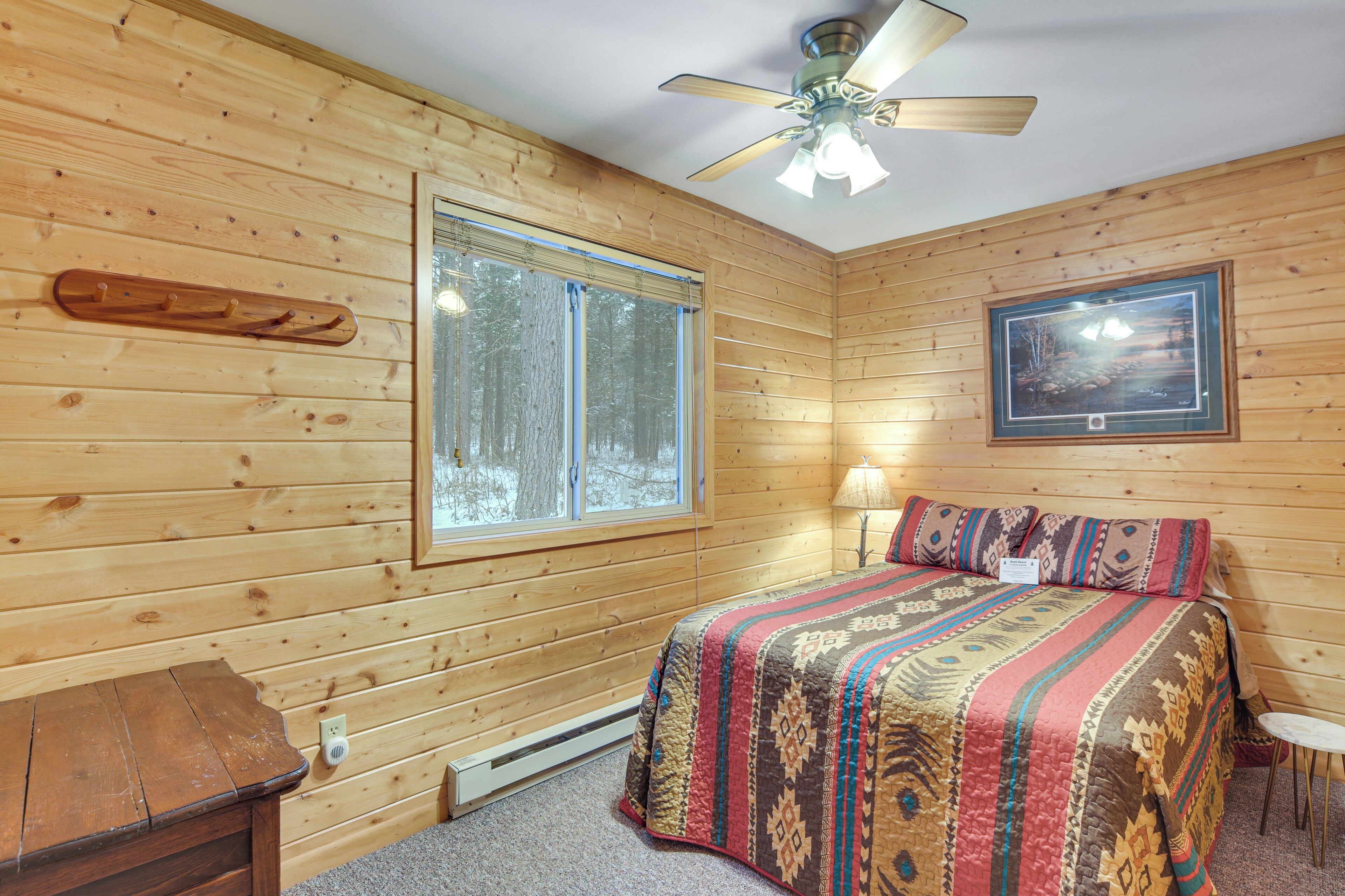 A wooden cabin room with a bed, a ceiling fan, a window, and a framed picture on the wall.