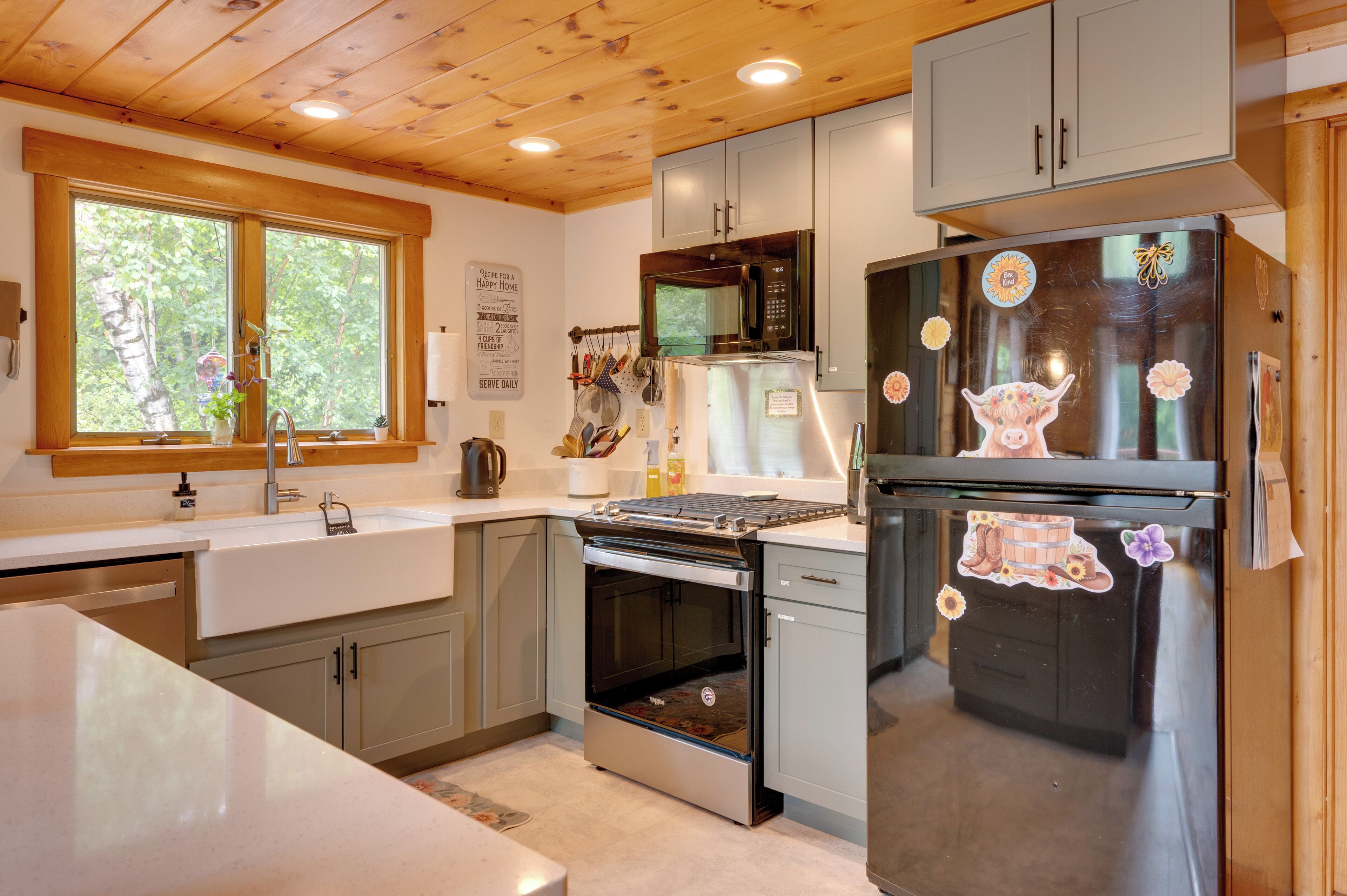 Una cocina con horno empotrado, microondas y refrigerador, techo de madera y ventana con vista a árboles.