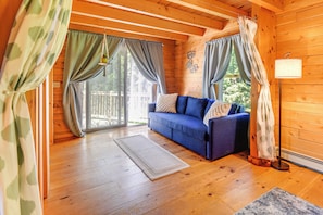 A cozy cabin interior with a blue sofa, wooden floors, and a large window with curtains. - Alexandria Cabin w/ Patio, Grill & Deck (Bristol)