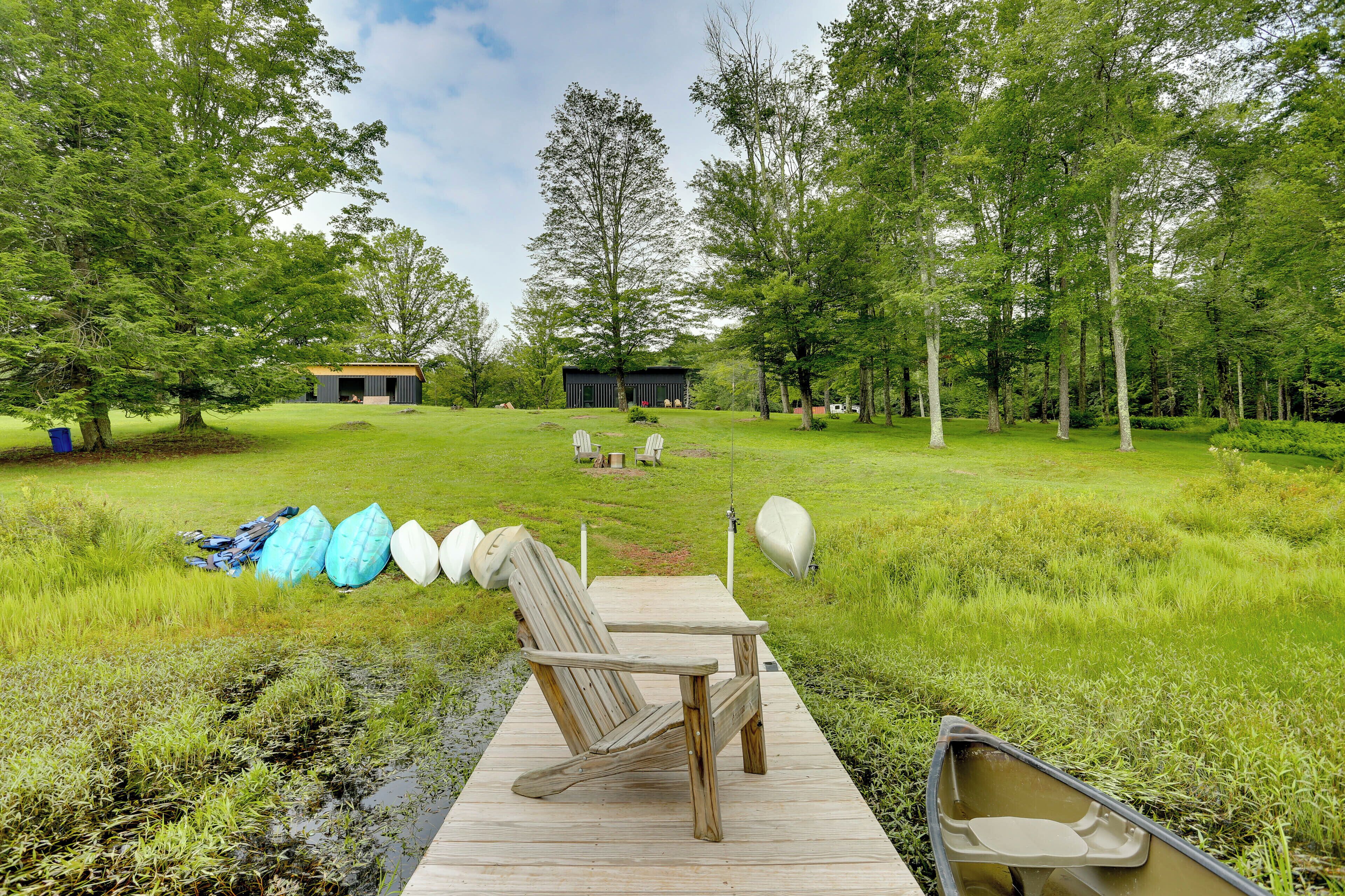 A wooden dock leading to a grassy area with a canoe and a rowboat, surrounded by trees and a small building in the background.