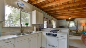 A kitchen with white cabinets, a stove, and a sink. There is a window with a view of trees and a clock on the wall. - Cozy Beachfront Cottage on Lake Ontario! (Henderson)