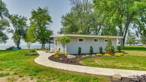 A single-story house with a curved driveway, surrounded by a well-maintained lawn and various shrubs. - Cozy Beachfront Cottage on Lake Ontario! (Henderson)