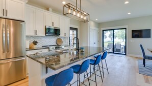 A modern kitchen with white cabinets, a stainless steel refrigerator, a microwave, and a large island with blue bar stools. - Spacious Lake Ontario Getaway: Steps to Beach! (Henderson)