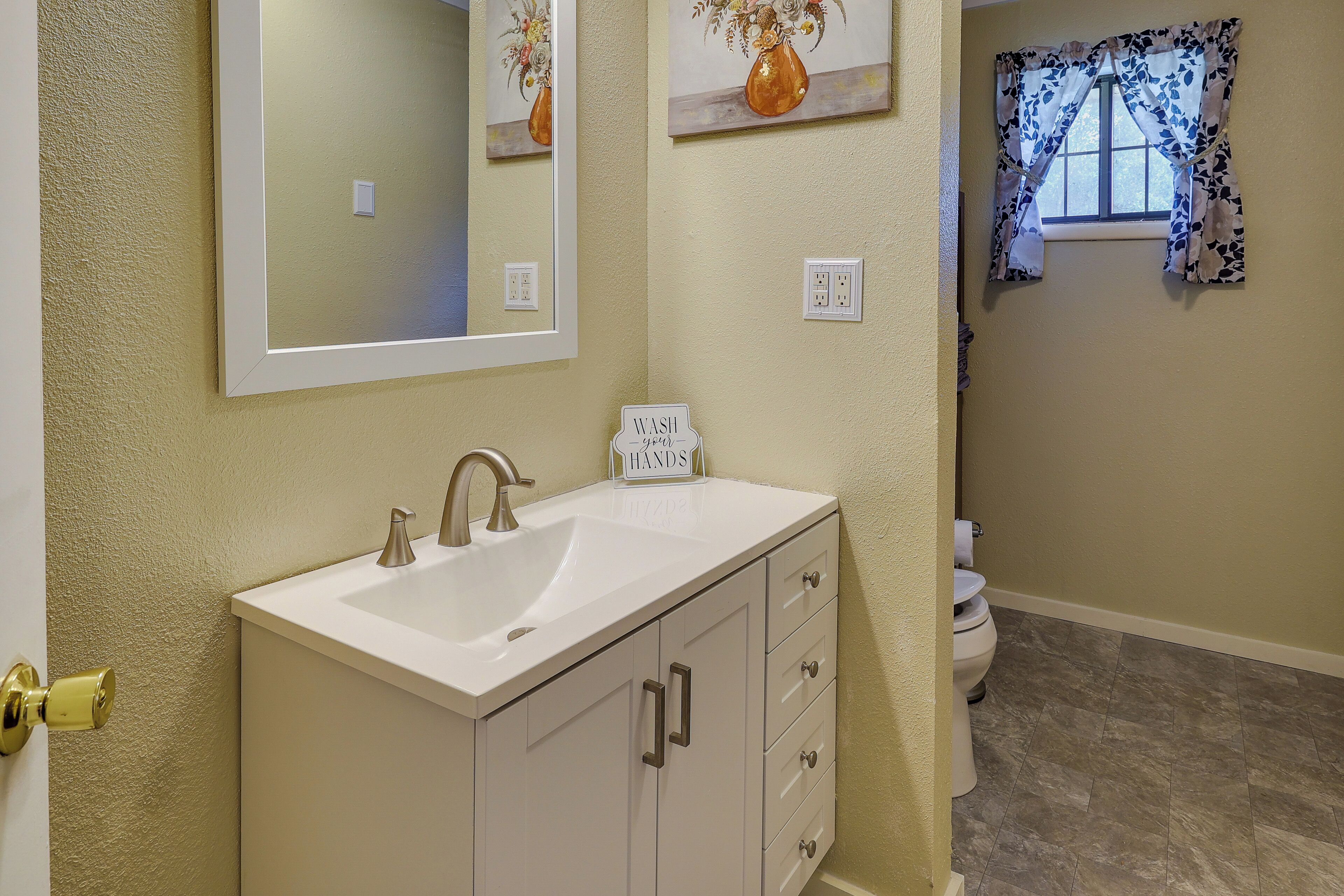 A bathroom with a white sink, mirror, and cabinet, a toilet, and a window with floral curtains.