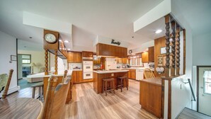 A modern kitchen with wooden cabinets, a central island with stools, and a clock on the wall. - Group-friendly Lake Erie Getaway w/ Private Yard (Conneaut)