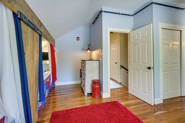 A hallway with a red rug, wooden flooring, a metal lamp, a red trash can, and a door with a white frame.