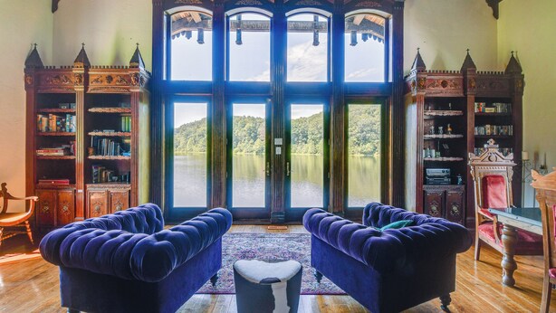 A room with two plush armchairs facing a large window with a view of a lake and hills. There are wooden bookshelves on either side of the window.