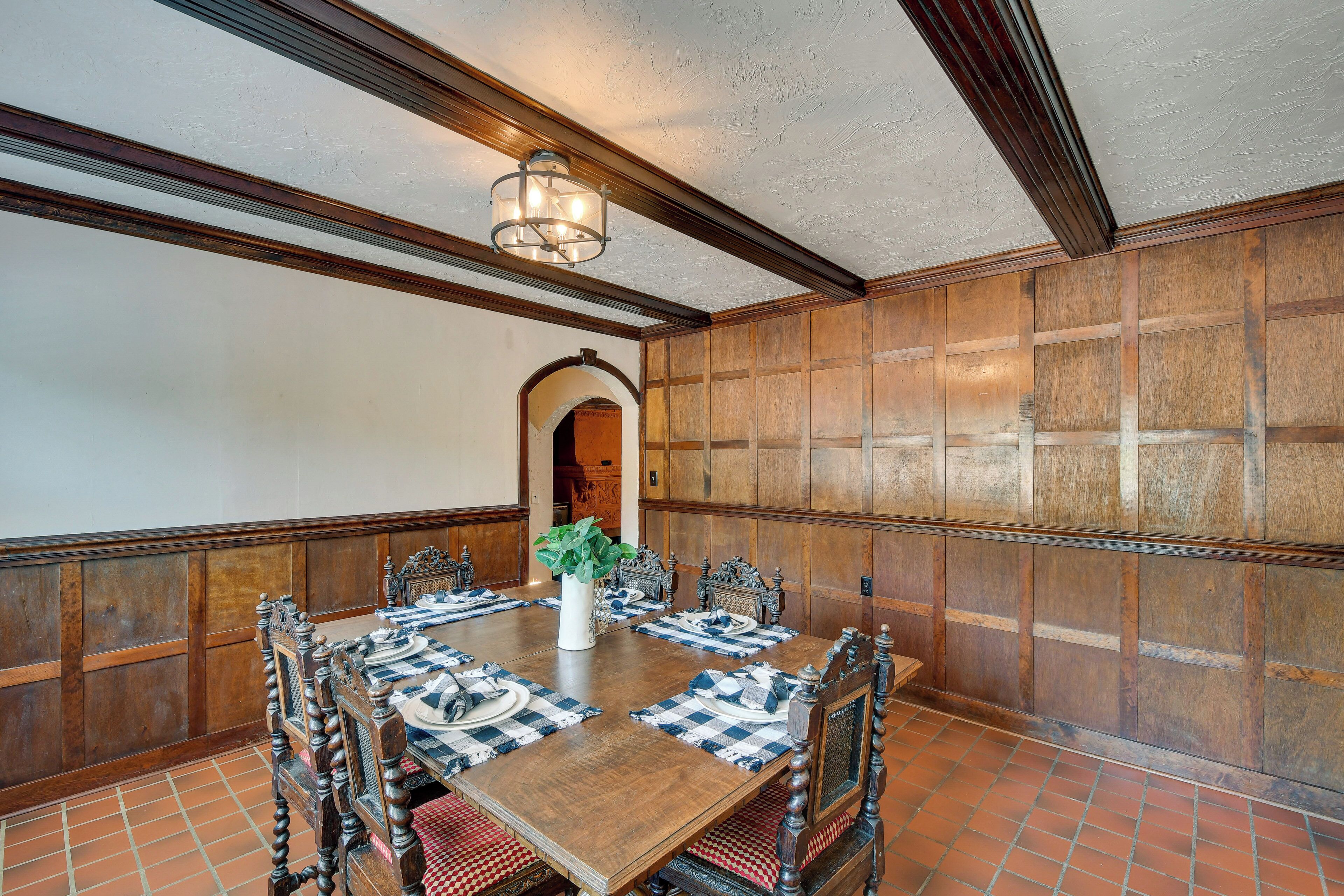 A dining room with a wooden table set for six, flanked by ornate chairs, a chandelier, and a large vase with a plant.