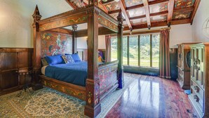 A four-poster bed with a blue bedspread, a wooden floor, and a large window with curtains.