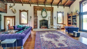 A spacious living room with a fireplace, plush purple sofas, a large patterned rug, and wooden beams on the ceiling.