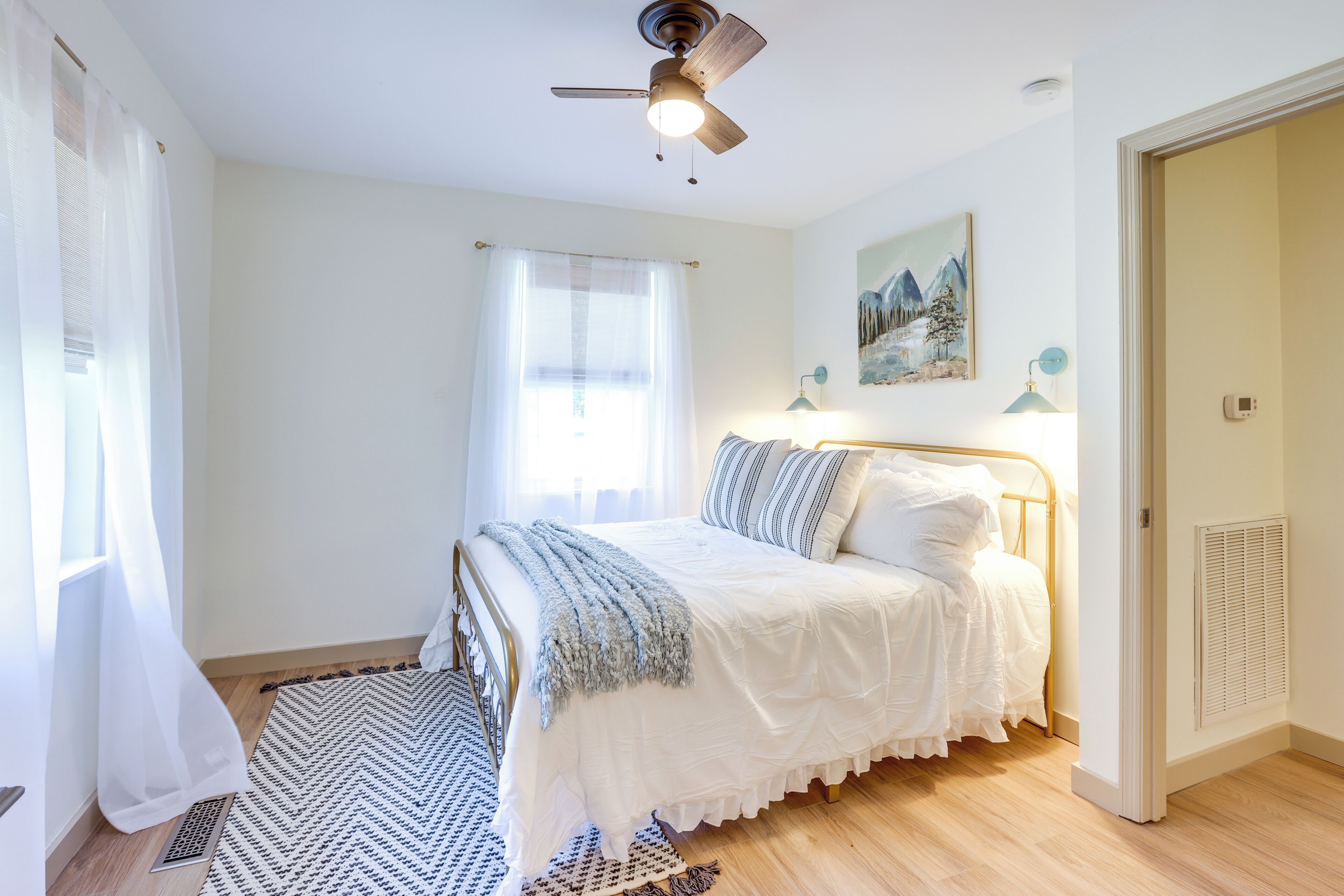 A bedroom with a bed, a ceiling fan, a window with sheer curtains, a painting on the wall, and a radiator.