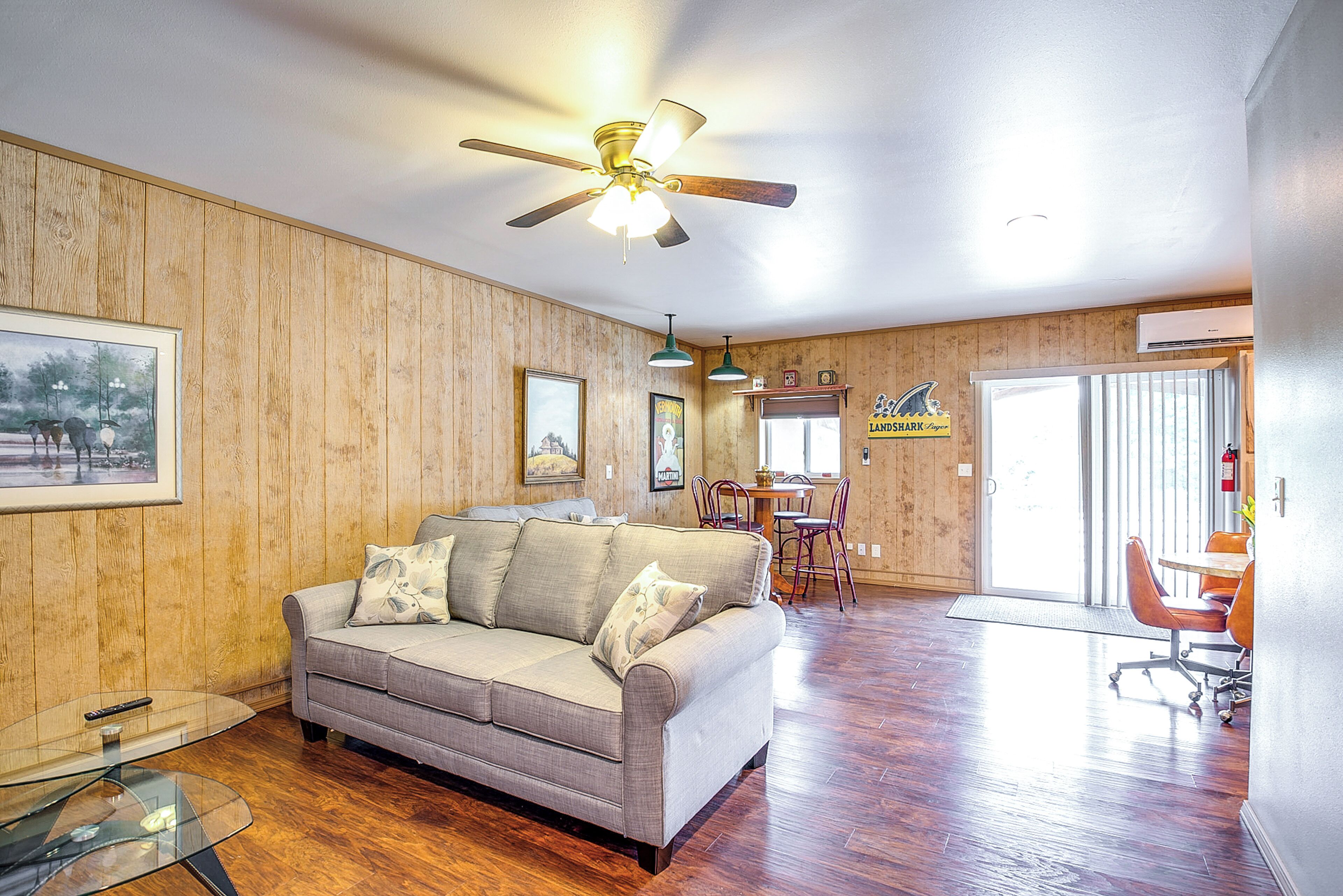 Una sala de estar con un revestimiento de madera, un ventilador de techo, un sofá, una mesa de centro de vidrio y una zona de comedor con una mesa y sillas.