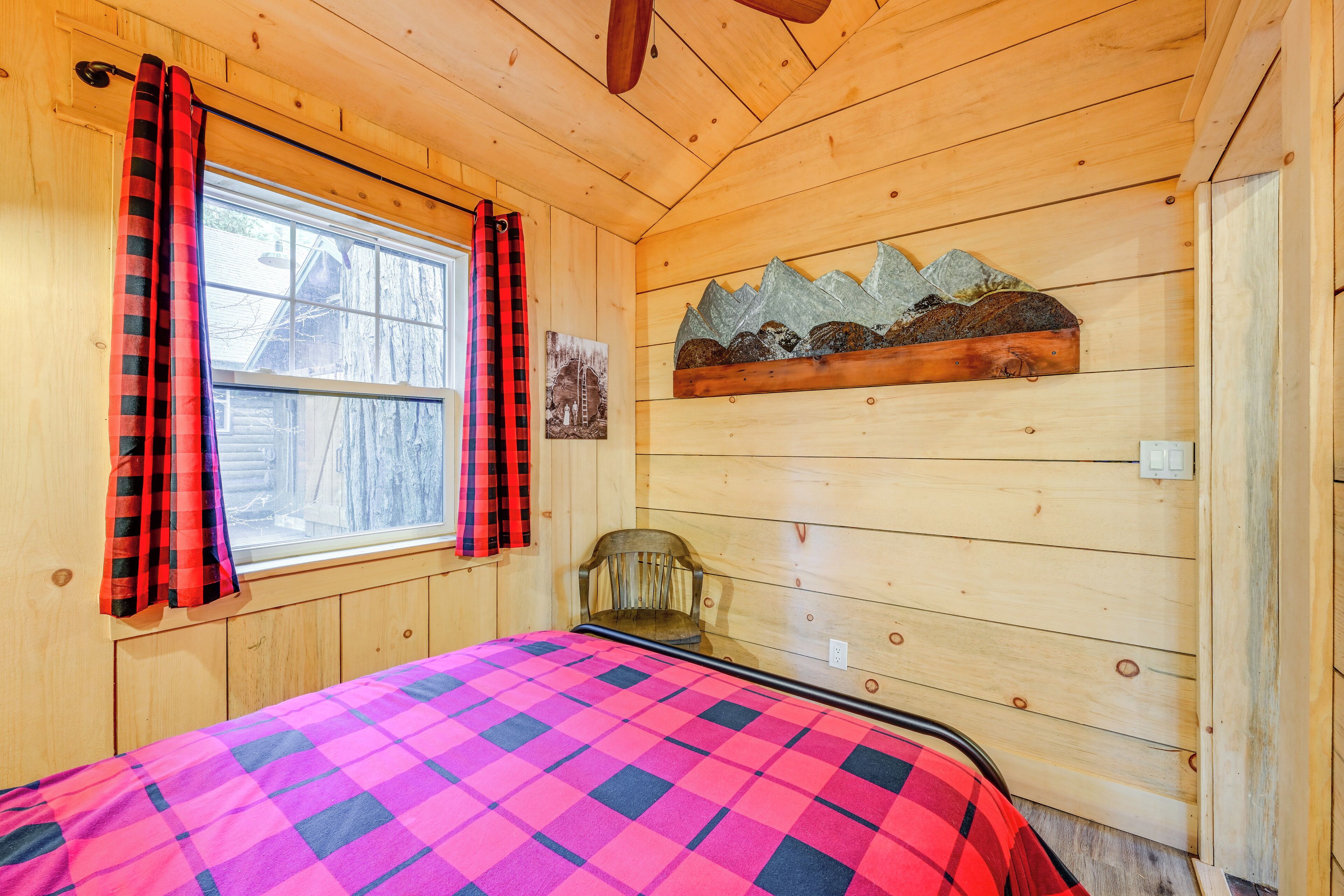 Un dormitorio de cabaña de madera con una cama, una silla, un ventanal con cortinas y un adorno en la pared.
