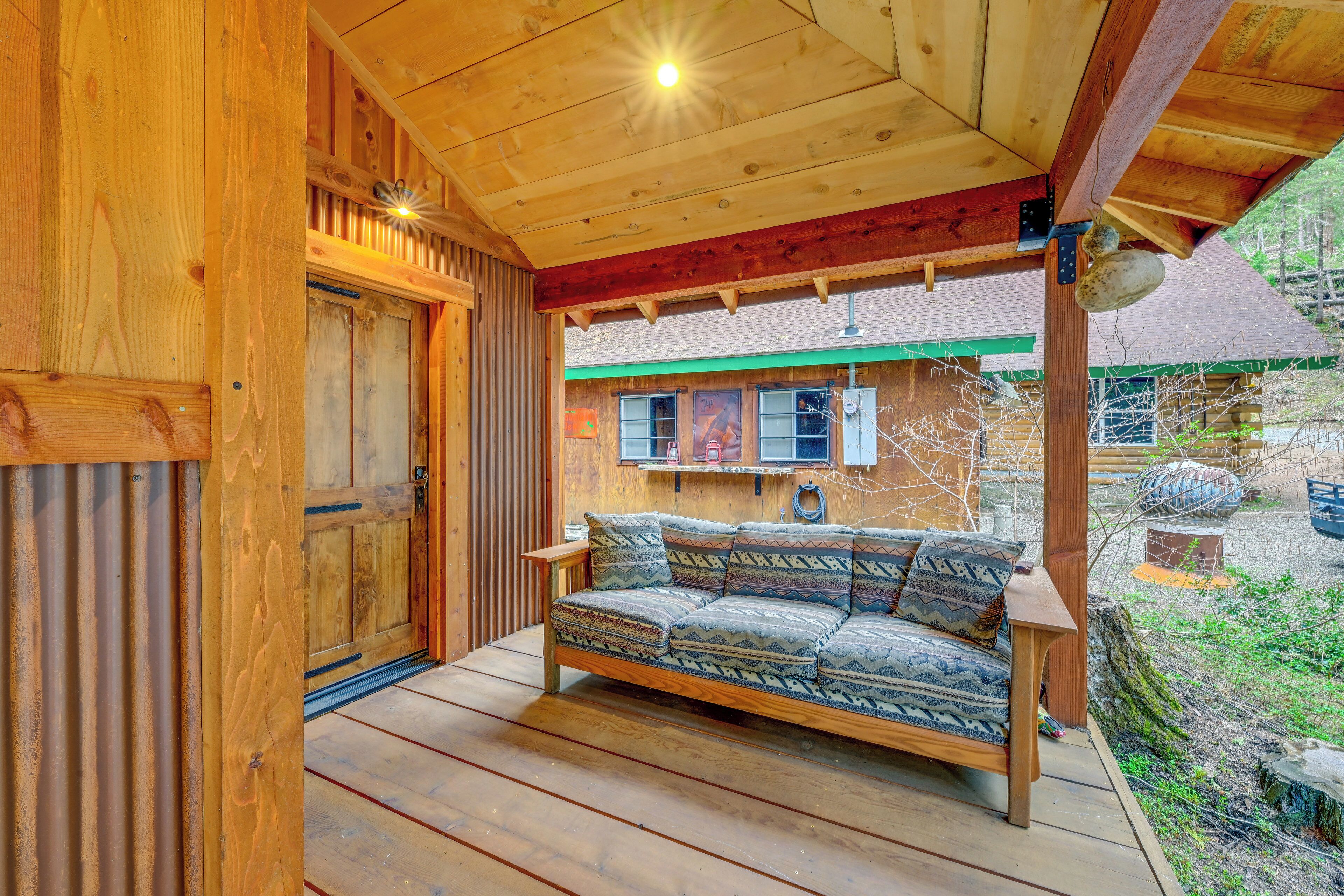 Una cabaña de madera con una terraza de madera, un sofá con cojines estampados y una vista de un edificio con techo verde al fondo.