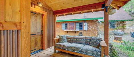 Una cabaña de madera con una terraza de madera, un sofá con cojines estampados y una vista de un edificio con techo verde al fondo.
