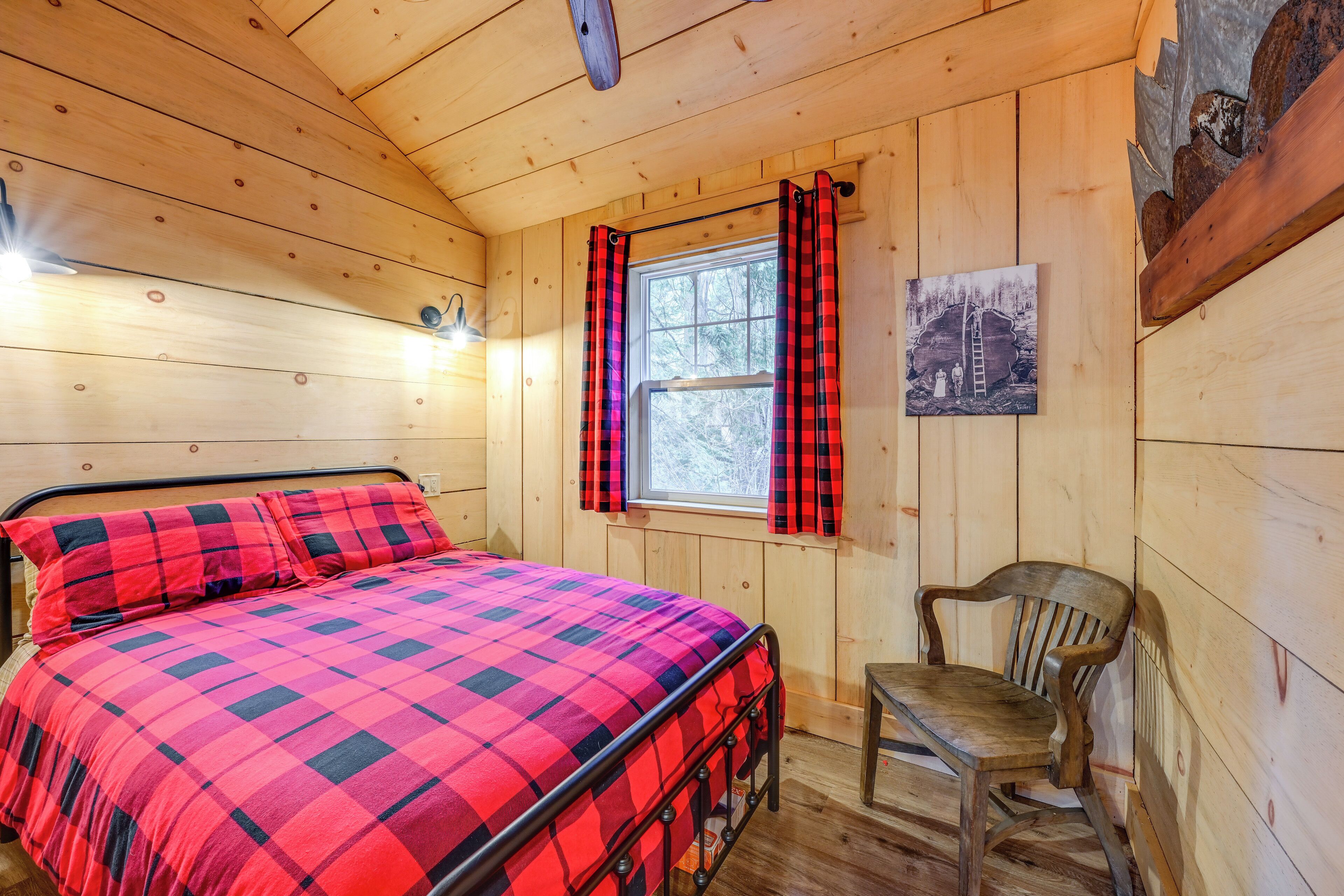 Una habitación de cabaña de madera con una cama, una silla, un ventanal con cortinas escocesas y un cuadro en la pared.