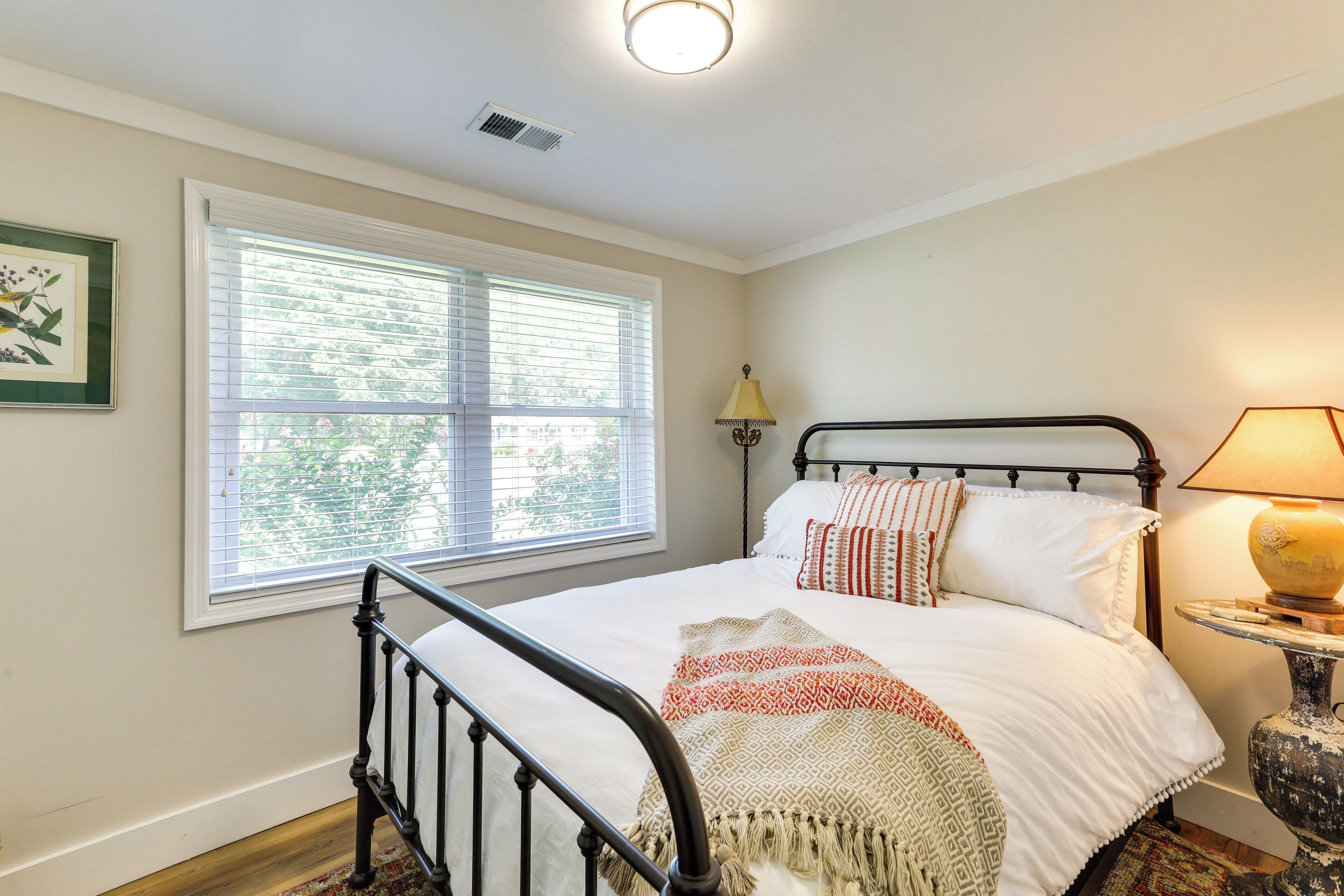 A bedroom with a bed, a window with blinds, a bedside lamp, and a framed picture on the wall.