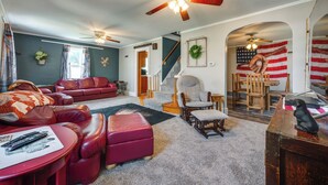 A living room with a red leather sofa, a red ottoman, a wooden coffee table, and a ceiling fan. There is a staircase leading to another level, and an American flag is visible on the wall. - Cozy Iowa Getaway w/ Patio & Fenced-in Yard! (Dyersville)