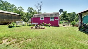 A red barn with a satellite dish, surrounded by a grassy yard with potted plants and a wooden bench. - Charming Oklahoma Retreat w/ Grill - Pets Welcome! (Spencerville)