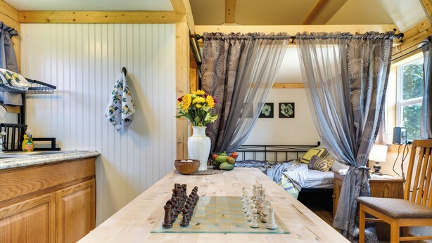 A wooden dining table with a chessboard in the center, a wooden bench, and a bed with a canopy in the background. - Charming Oklahoma Retreat w/ Grill - Pets Welcome! (Spencerville)
