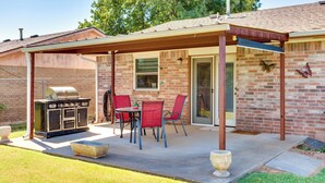 A covered patio with a barbecue grill, outdoor seating, and a brick house.