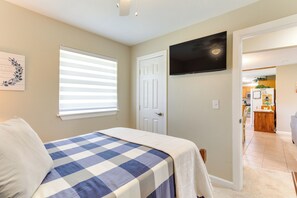 A bedroom with a checkered bedspread, a television mounted on the wall, a door leading to a kitchen, and a window with blinds. - Cozy Oklahoma Retreat w/ Covered Patio & Gas Grill (Mustang)