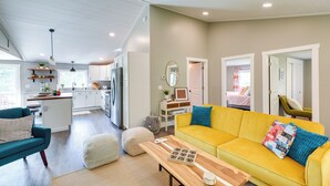 Un espace de vie moderne avec un canapé jaune, un fauteuil turquoise et une table basse en bois. On aperçoit à l’arrière-plan une cuisine avec des meubles blancs et un réfrigérateur.