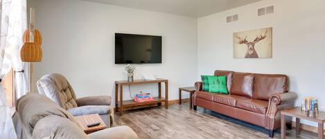 A living room with a brown leather sofa, beige armchairs, a wooden coffee table, and a flat-screen TV mounted on the wall.
