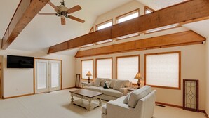 A spacious living room with a high ceiling, wooden beams, a ceiling fan, a large window, a television, and a fireplace. - Gordonsville Retreat: Deck w/ Spectacular Views! (Gordonsville)