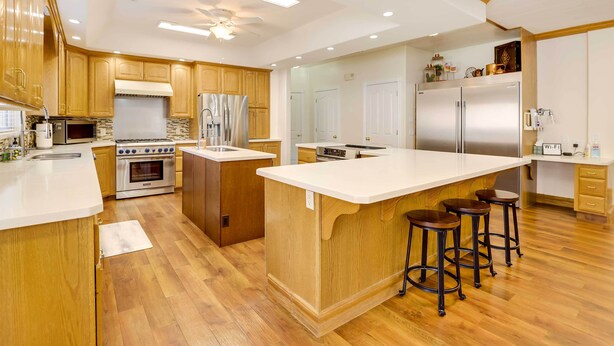A modern kitchen with wooden cabinets, a large island with stools, stainless steel appliances, and a ceiling fan. - Spacious Hildale House w/ Large Deck! (Hildale)