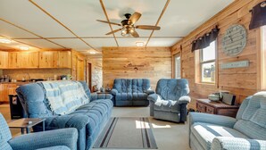 Ein gemütliches Blockhaus-Interieur mit einem Wohnzimmer, das blaue Polstermöbel, einen Deckenventilator, Holzvertäfelung und eine Küchenzeile zeigt.