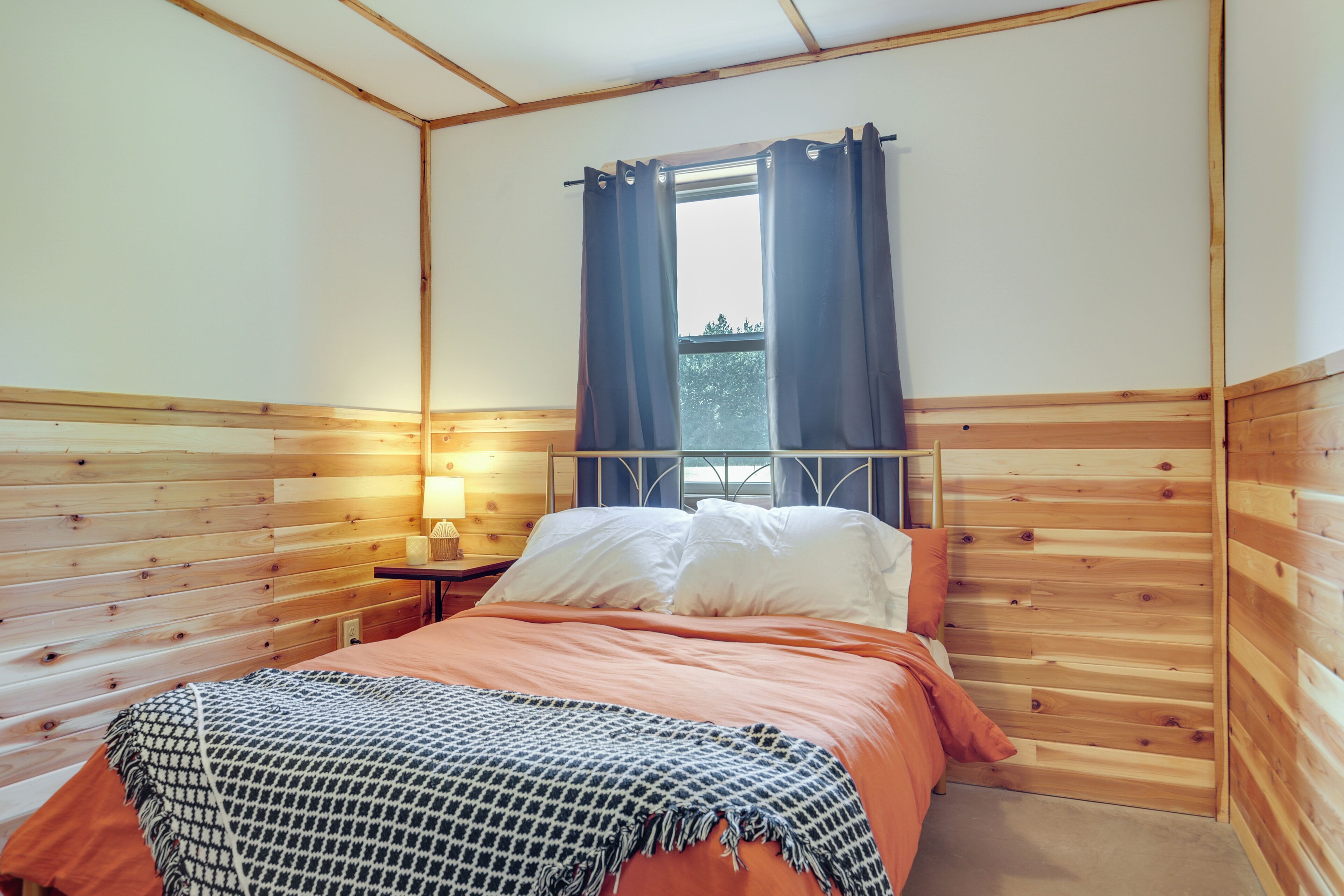 A bedroom with wooden paneling, a bed with white and orange bedding, a nightstand with a lamp, and a window with curtains.