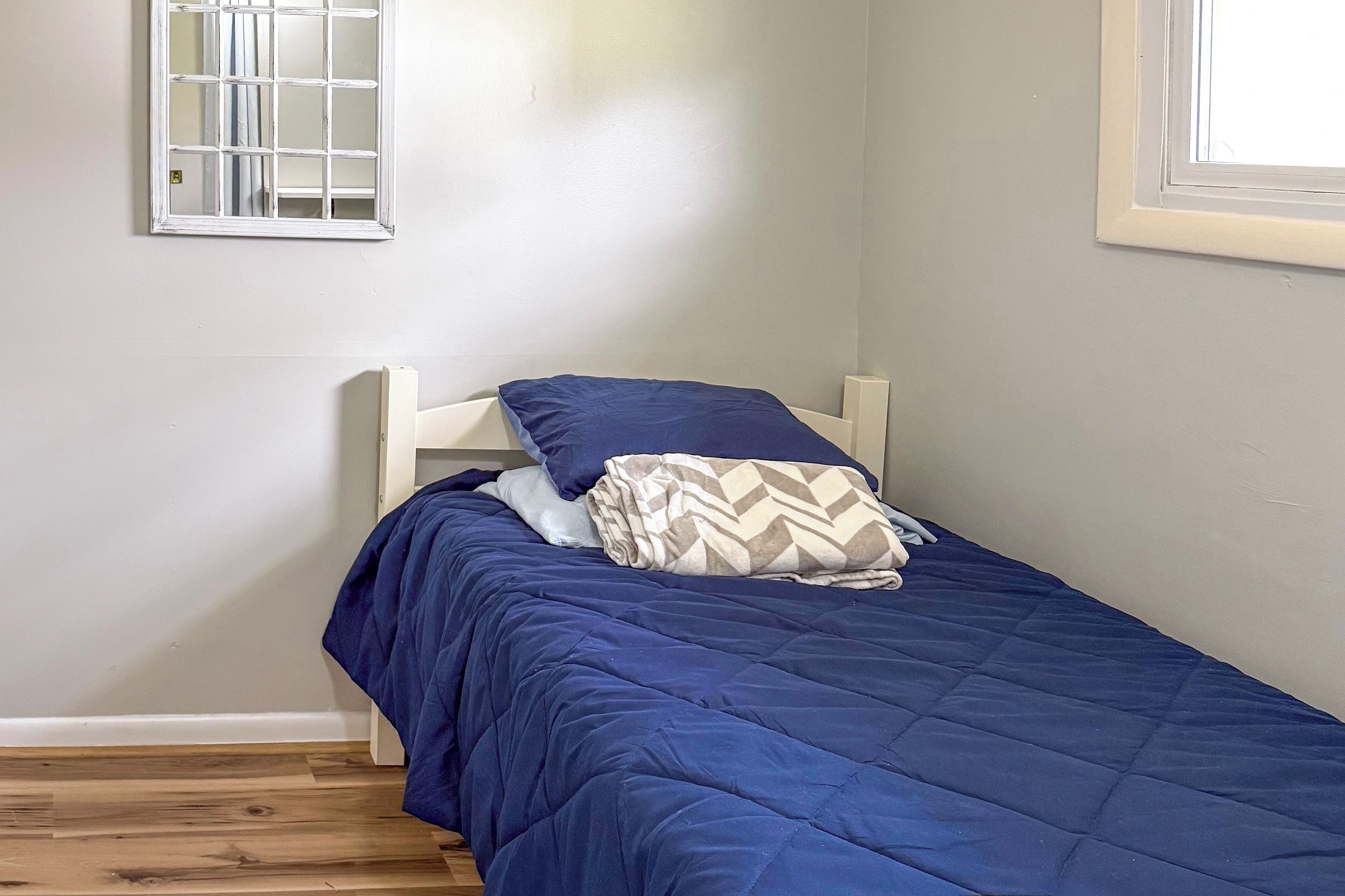 Een kleine kamer met een enkel bed, een houten vloer, een wit hoofdeinde en een raam met een rasterpatroon.
