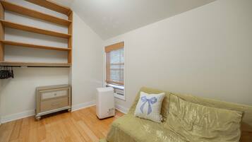 A room with a wooden wardrobe, a white radiator, a beige sofa with a decorative pillow, and a window with blinds.