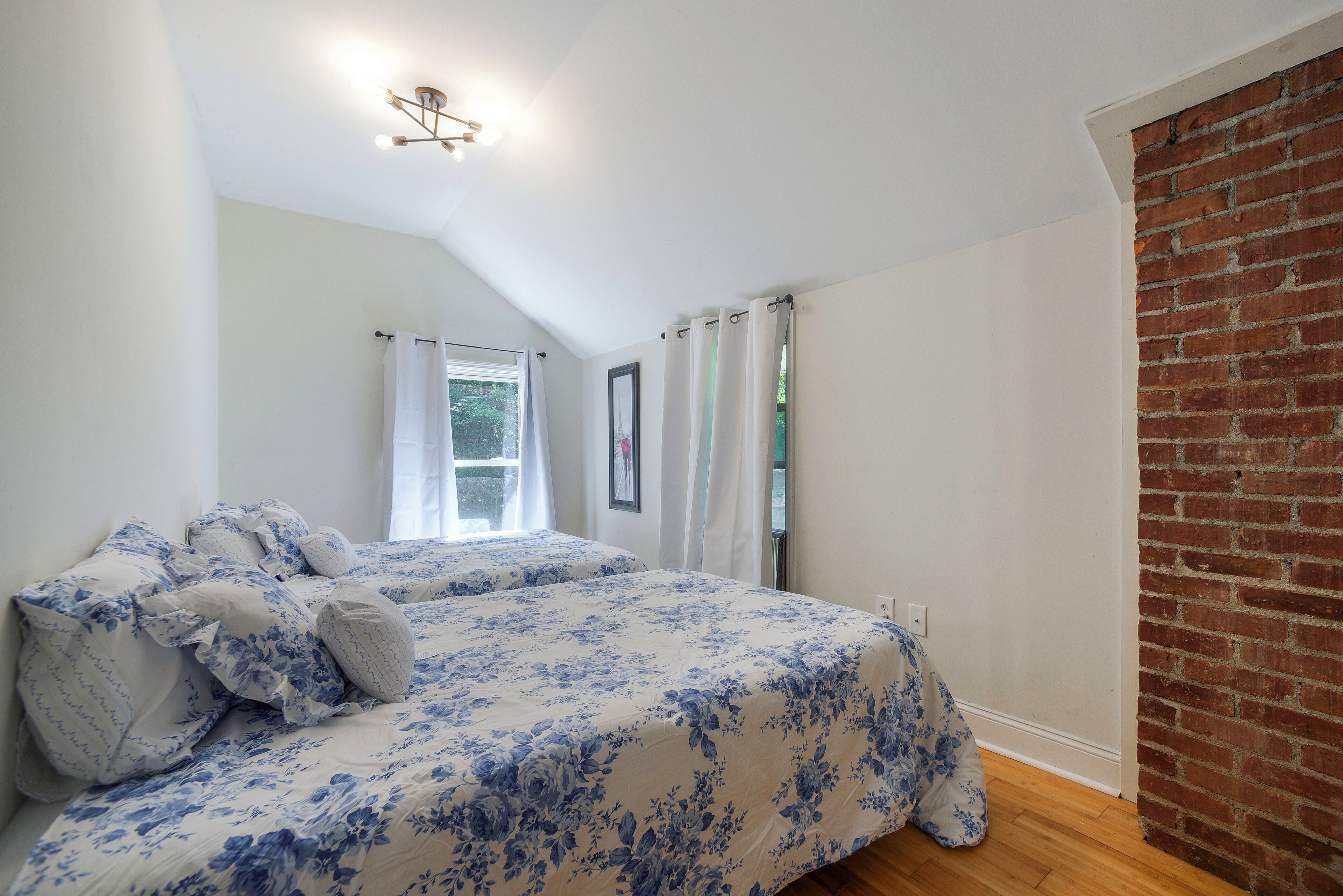 Ein Schlafzimmer mit Bett, einem Fenster mit Vorhängen, einer Ziegelwand und einer Deckenleuchte.