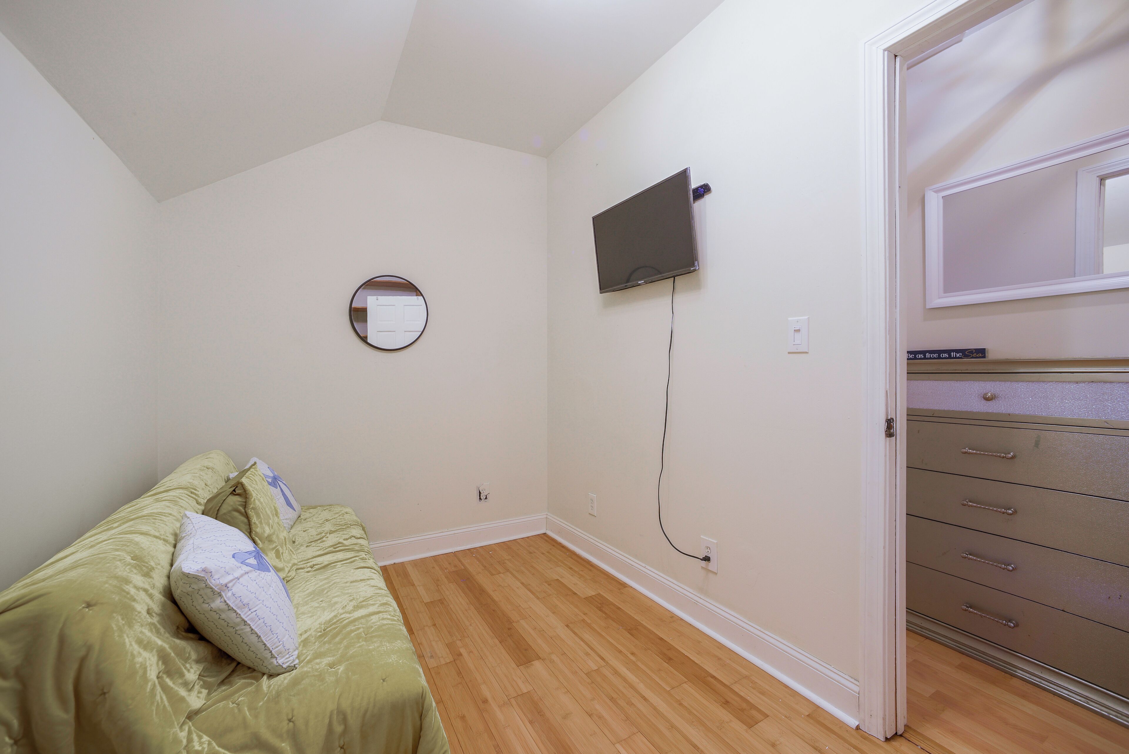 A bedroom with a bed, a round mirror, a flat-screen TV mounted on the wall, and a dresser with drawers.