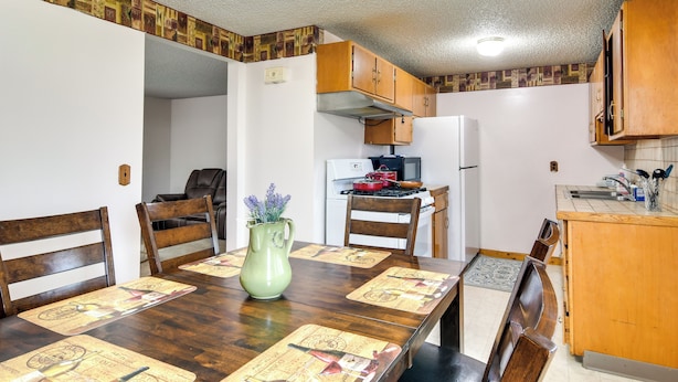 Un coin repas avec une table en bois dressée pour quatre personnes, un pichet vert orné de fleurs et, au fond, une cuisine.