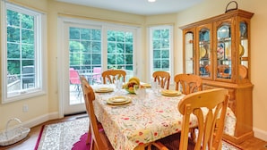A dining room with a table set for four, surrounded by chairs, a wooden cabinet with glass doors, and a view through large windows. - Charming Flushing Cottage w/ Deck + Spacious Yard! (Flushing)