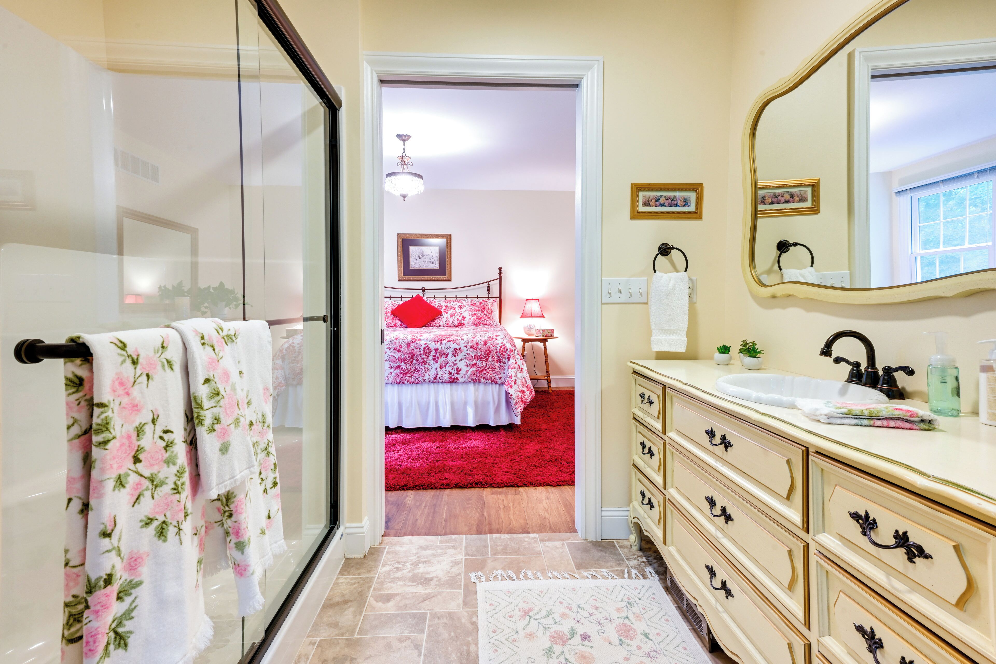 A bathroom with a glass shower door, a sink with a mirror, and a red carpet leading to a bedroom.