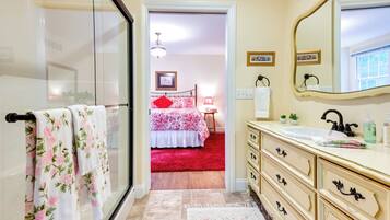 A bathroom with a glass shower door, a sink with a mirror, and a red carpet leading to a bedroom.