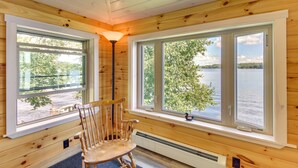Una cabaña con paredes de madera, un ventanal con vistas a un lago, una silla de madera y una lámpara de pie.