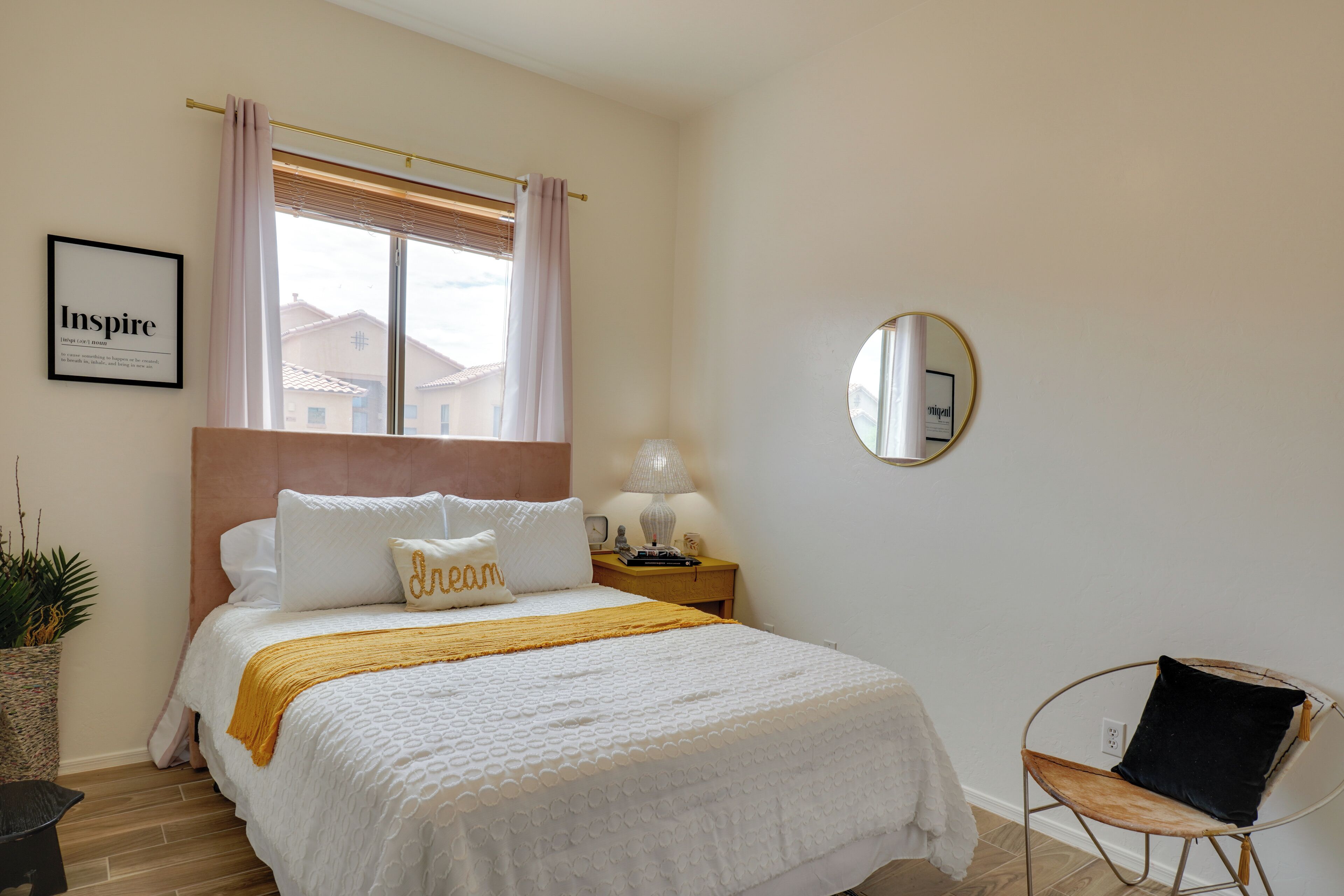 A bedroom with a bed, a bedside table, a lamp, a chair, a window with curtains, a framed picture on the wall, and a potted plant.