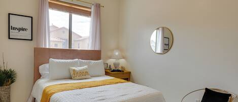 A bedroom with a bed, a bedside table, a lamp, a chair, a window with curtains, a framed picture on the wall, and a potted plant.
