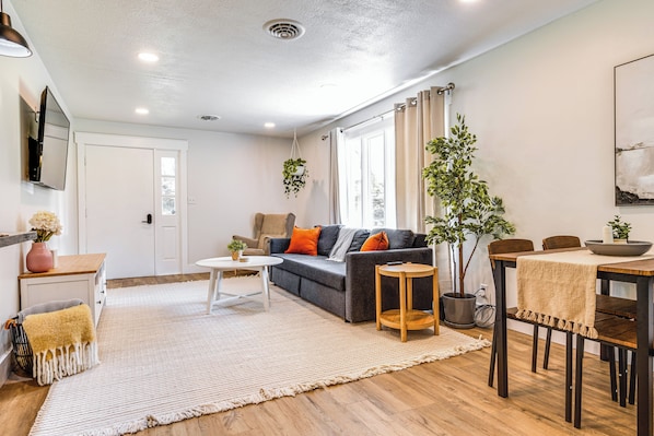 A modern living room with a sofa, coffee table, and dining table. There is a large window with curtains, a potted plant, and a flat-screen TV mounted on the wall.