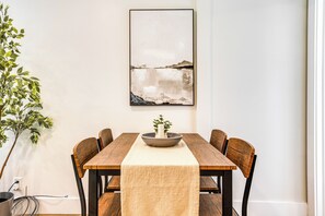 A dining table with a woven table runner, surrounded by wooden chairs, and a framed abstract painting on the wall.