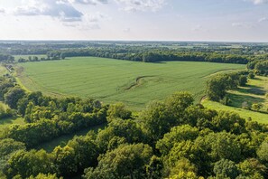 An aerial view of a large green field with a winding path, surrounded by dense trees and open fields.