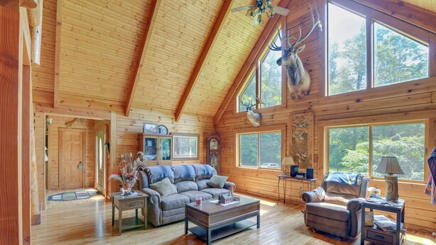 A spacious log cabin with a high ceiling, wooden floors, a large sofa, a coffee table, a fireplace, and large windows offering views of the surrounding forest. - Serene Valley Bend Cabin Rental: 7 Private Acres! (Beverly)