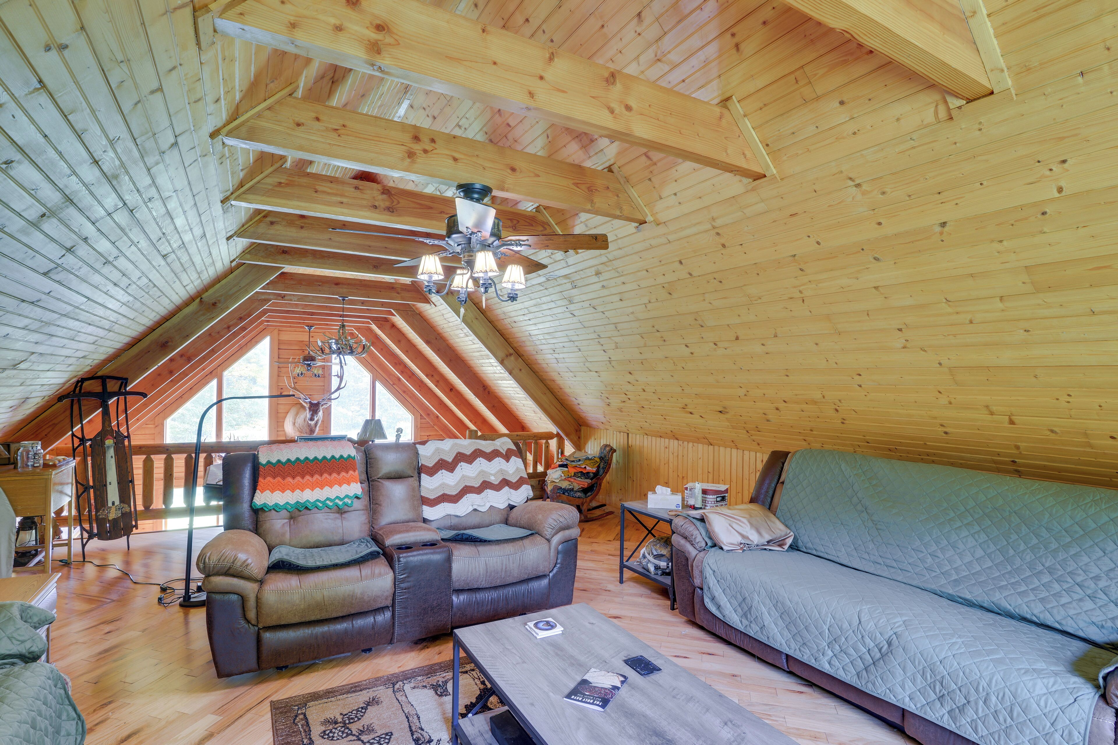 A cozy attic room with a wooden ceiling, a ceiling fan, a sofa, a chair, and a table.