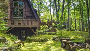 Un chalet rustique doté d’une terrasse en bois, entouré d’une zone boisée avec un foyer et des murs en pierre.