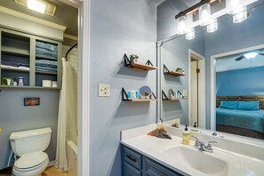 Un baño con inodoro blanco, un lavabo con espejo y repisas con artículos de aseo.